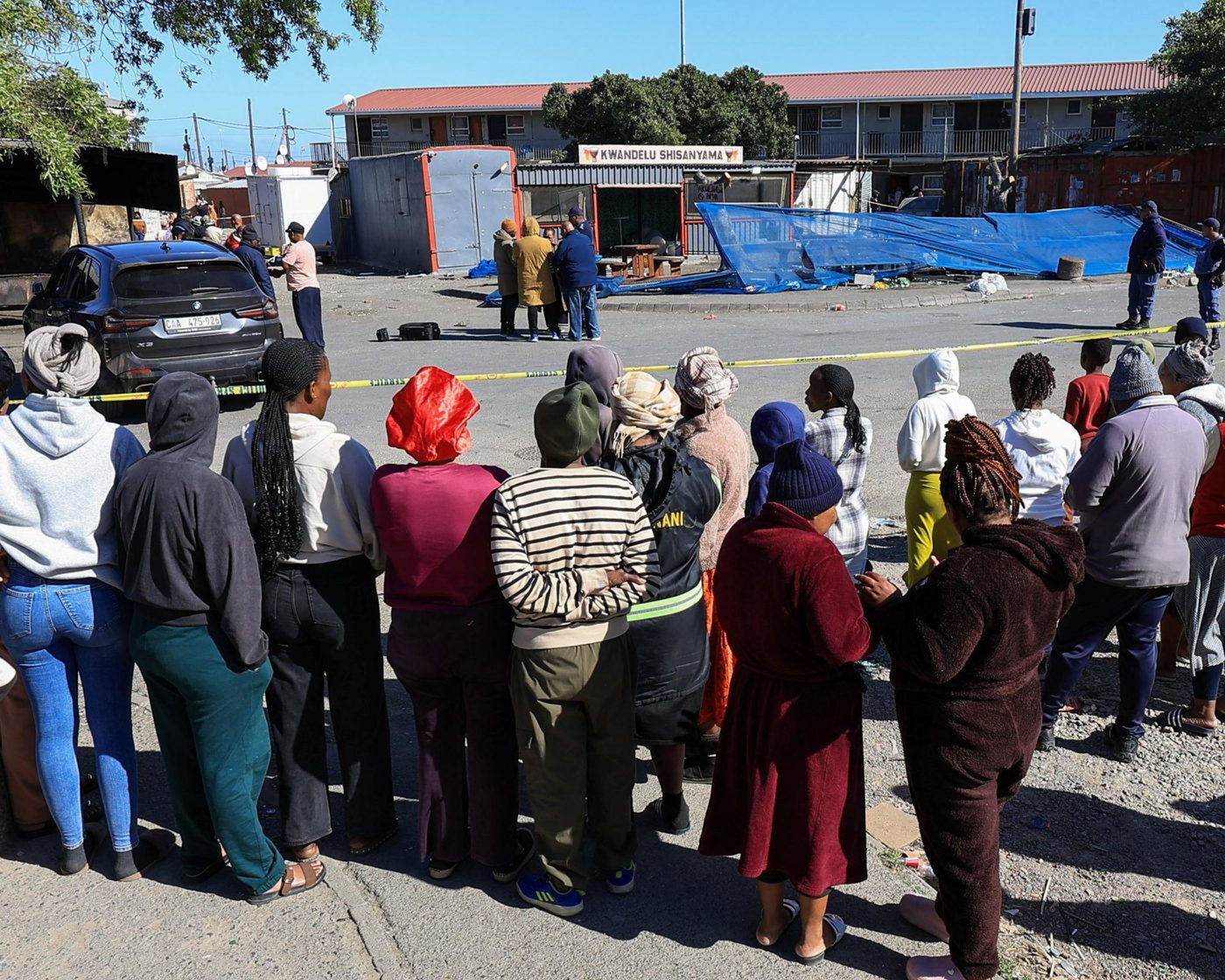 Archive documentary photograph of the KwaNdelu Shisanyama shooting scene in Lwandle, September 2026, with residents gathered behind police tape
